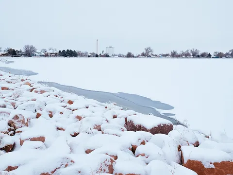 Windsor Lake frozen and snow-covered in winter, red sandstone riprap on the near shore and the town’s houses, bare trees and grain elevator along the far one