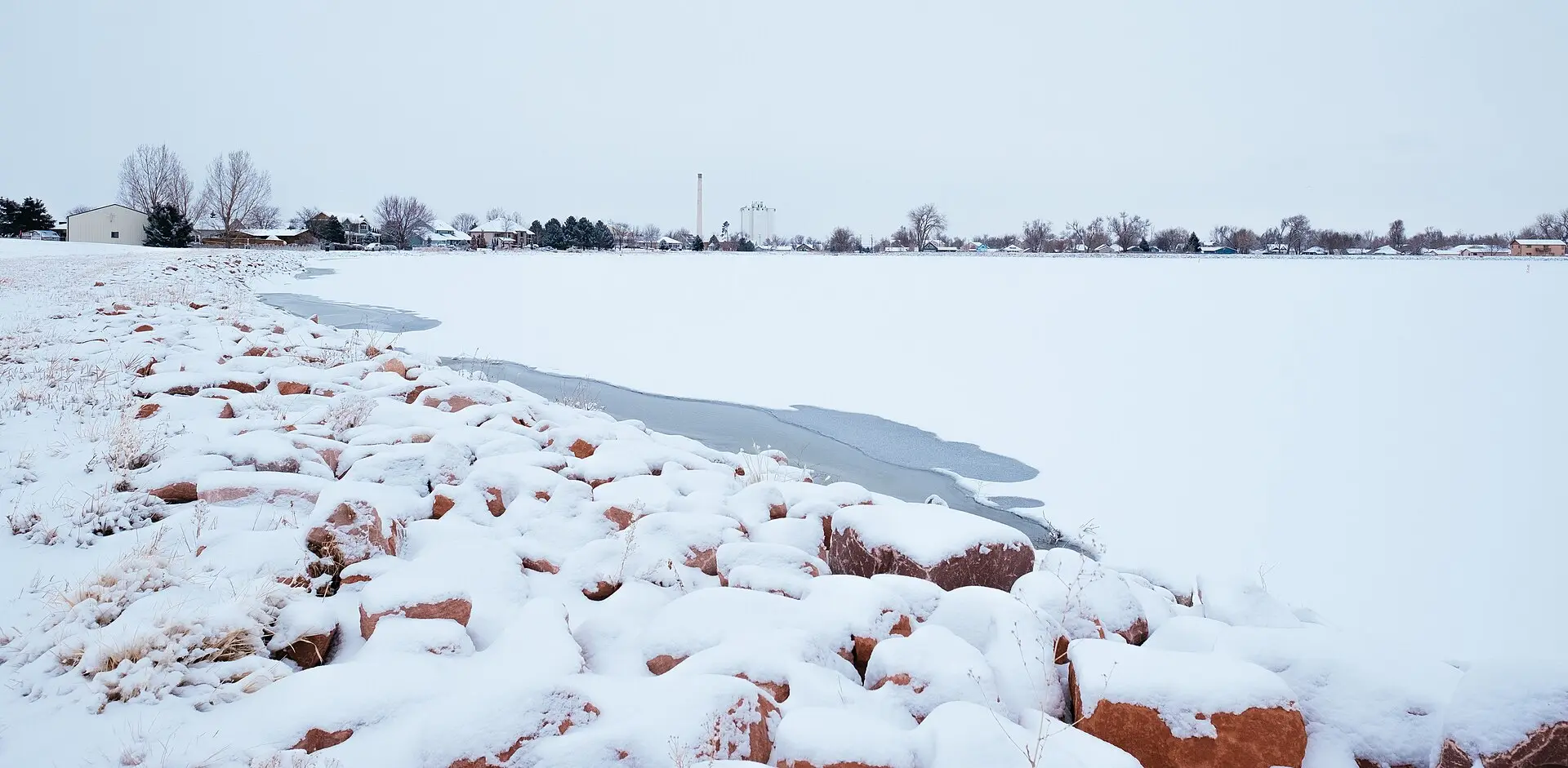 Windsor Lake frozen and snow-covered in winter, red sandstone riprap on the near shore and the town’s houses, bare trees and grain elevator along the far one