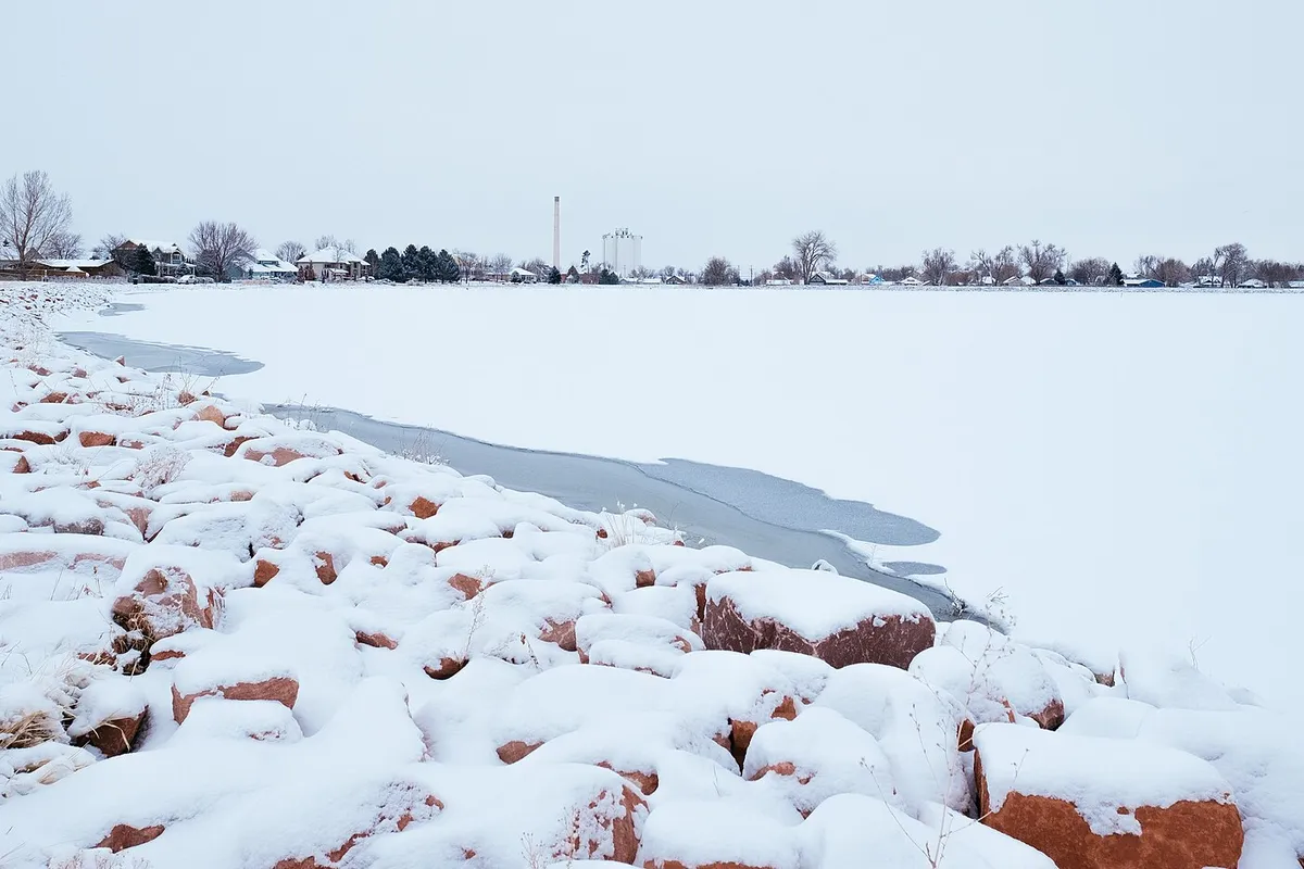 Windsor Lake frozen and snow-covered in winter from the trail on the near shore, red sandstone riprap at the water’s edge and the town’s houses, bare trees and grain elevator along the far one